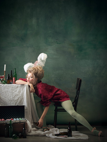 Drunk with empty bottles. Young woman as Marie Antoinette isolated on dark green background. Retro style, comparison of eras concept. Beautiful female model like classic historical character, old-fashioned.