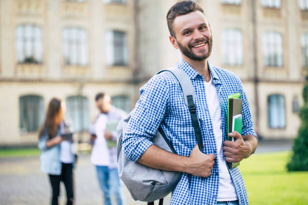 istock Young handsome bearded student guy in casual clothes with backpack and books in hands is walking and posing on a university  buildings background 1266797311