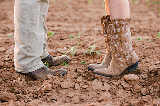 Male and female couple wearing cowboy boots in a dirt field. The woman is tiptoeing.
