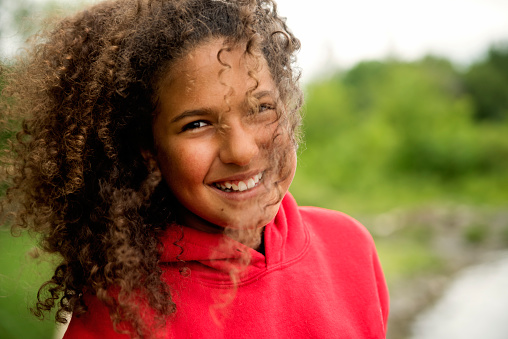 Mixed-race teenage girl portrait on riverside. She is wearing a red hoodie, have very curly hair and is looking at the camera with a smile. Wind in hair. Horizontal head shot outdoors with copy space.