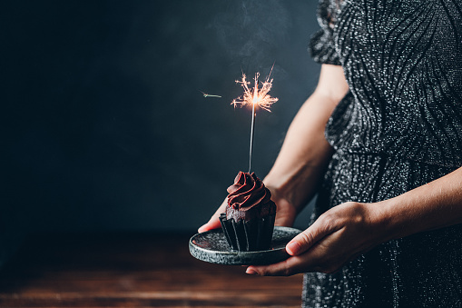 Cropped shot of a woman holding chocolate cupcake with burning sparkler. Female hand holding delicious birthday cupcake with firework candle