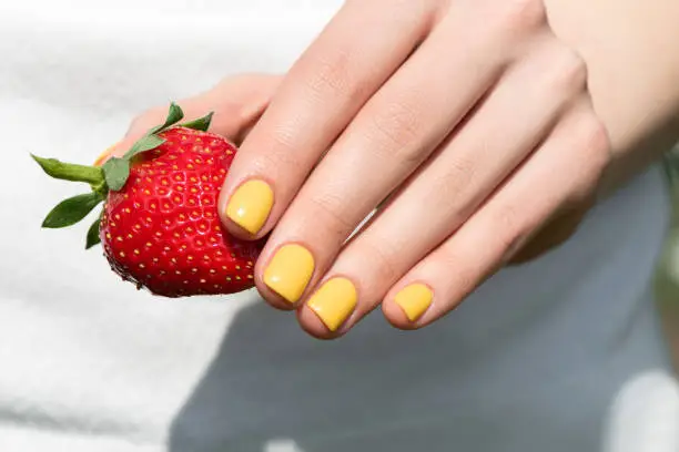 Photo of Close up of female hand with pretty yellow nail design manicure holding ripe strawberry.