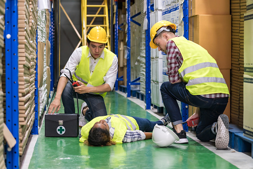 Warehouse worker do first aid to his colleague lying down on warehouse floor after accident while working. Using for industrial safety first and business insurance concept.