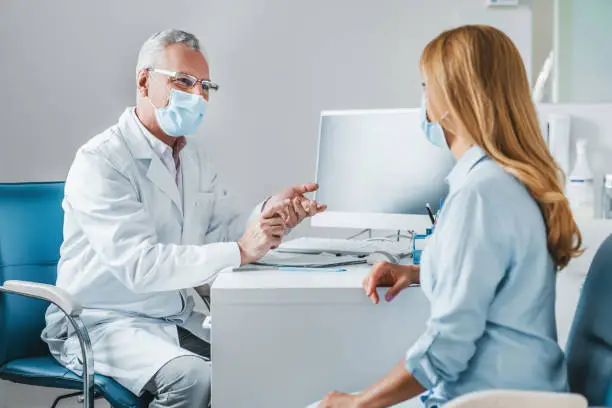 Photo of Senior male doctor wear protection face mask talking with patient in clinic office