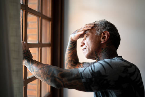 homme mûr inquiet restant à la maison, près d’une fenêtre, avec la main dans la tête - tender emotion photos et images de collection