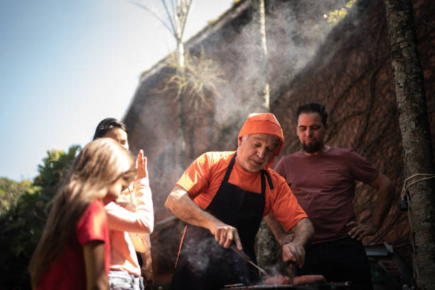 grand-père coupant des tranches de viande pendant un barbecue avec la famille - barbecue grill chef barbecue sausage photos et images de collection