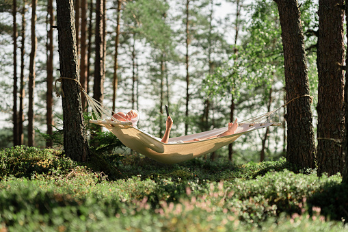 A man is lying in a hammock in the sun, he is reading a book on his digital tablet. The hammock is hanging in a beautiful green forest in Sweden.