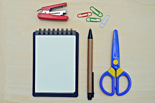 A horizontal photograph of a  blank open white page of a spiral notepad, along with stationery items over a wooden background. Suitable to use as school or office supplies related backdrops. There is a red stapler, a ballpoint pen, a blue coloured scissors and four paper clips.