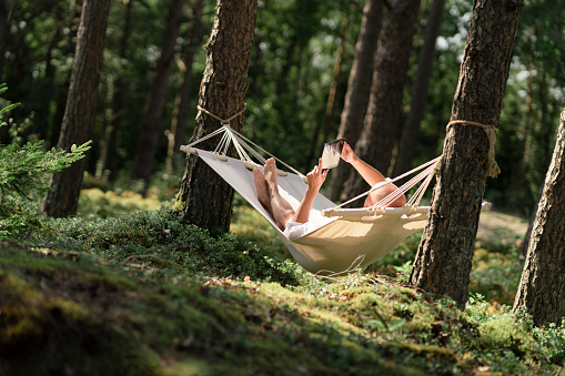 A man is lying in a hammock in the sun, he is reading a book on his digital tablet. The hammock is hanging in a beautiful green forest in Sweden.