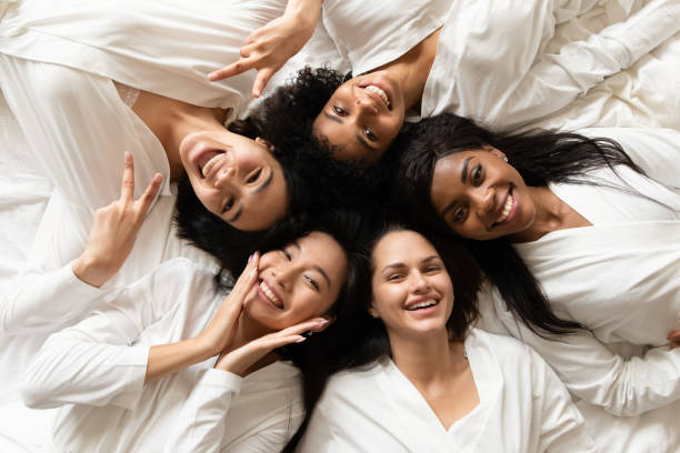 Diverse women lying in bed feels happy after spa procedures Top view five diverse women in white bathrobes lying in bed smile look at camera feels happy after body treatment, day spa procedures, resort beauty salon satisfied clients, bachelorette party concept bathrobe photos stock pictures, royalty-free photos & images