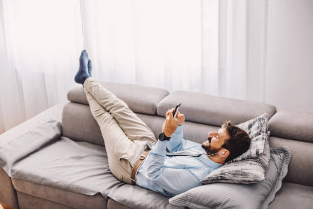 istock Young attractive bearded employee lying down on the sofa and using mobile phone during quarantine. Covid outbreak concept. 1266825886
