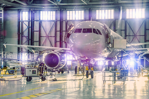 Civil airplane jet on maintenance of engine and fuselage check repair in airport hangar. Bright light purple tint at the gate.
