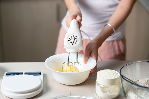Woman using electric mixer to mix ingredients for dough of sugar, beaten eggs, butter and flour while making cookies in the kitchen at home closeup