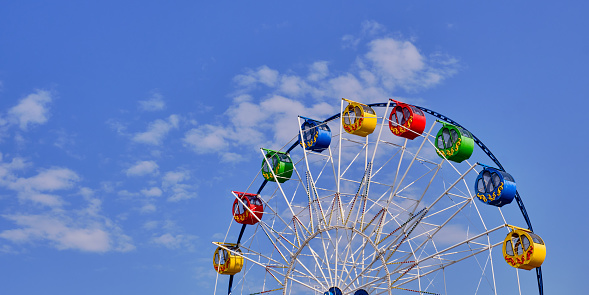 A colorful ferris wheel with round cabins decorated with ornaments against a blue sky with clouds. Copy space. Banner