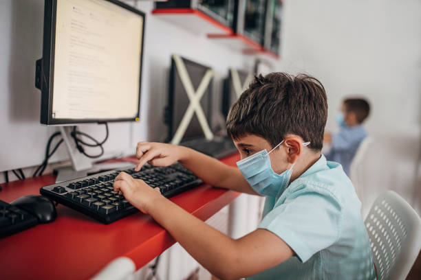 joven niño en edad preescolar que usa máscara protectora en el aula de computación - computer lab child internet development fotografías e imágenes de stock