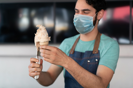 A young ice-cream seller is wearing protective face mask and preparing an ice-cream cone. COVID-19 concepts.