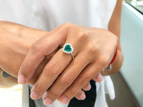 Bright green emerald heart shape with diamonds white gold ring on young woman’s hand close up