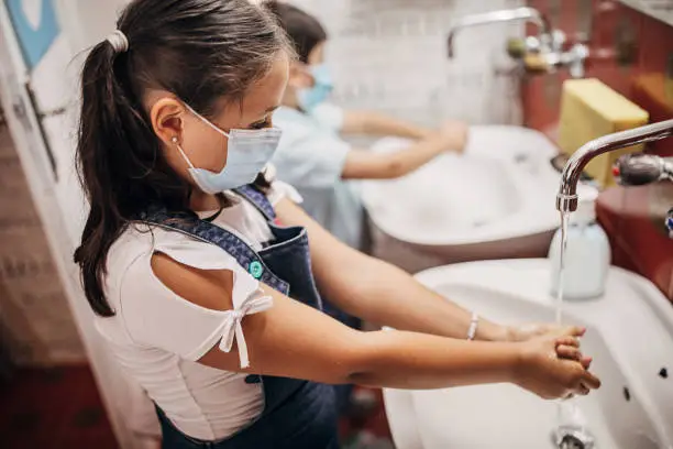 Photo of Young girl with protective face masks washing hands with soap in bathroom before they enter in classroom