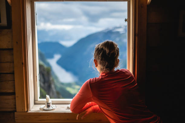 woman overlooking fjord in norway through window. - home interior cabin shack european alps imagens e fotografias de stock
