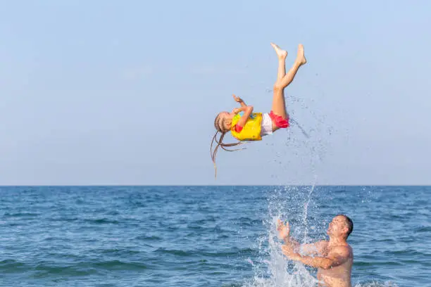 A strong Caucasian man in sea tosses a 4-5 year girl. A child in a life jacket with splashes flies upward, does a somersault, a coup, an acrabatic trick against the background of blue sky and sea.