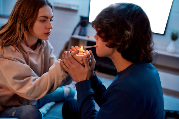 Young girl lighting cannabis cigarette for her boyfriend while sitting on the couch at home. Young couple smoking marijuana joint and relaxing Young girl lighting cannabis cigarette for her boyfriend while sitting on the couch at home. Young couple smoking marijuana joint and relaxing. Marijuana, drugs, cannabis and weed legalization blunts stock pictures, royalty-free photos & images