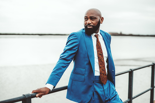 A handsome robust bald mature black guy with a beard and in an elegant blue costume with a tie is leaning against a railing of a metal fence on the pier and pensively looking aside