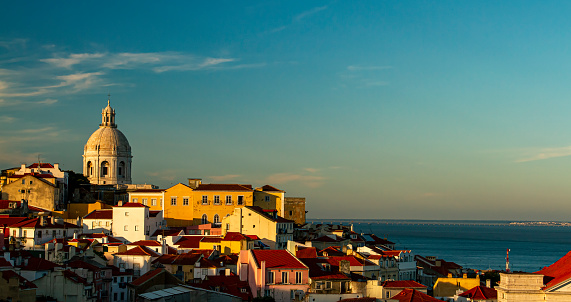 A sunset view of the historic Alfama district of Lisbon, Portugal. Image features the National Pantheon (the white dome overlooking the city), houses, Tagus River and Vasco da Gama bridge.