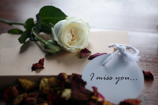 I miss you. Romantic note message written on white paper tag label with background of white rose lying  and dried petals scattered on book gift on messy table.