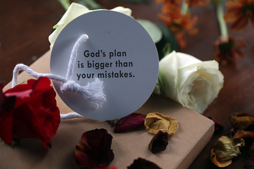 Inspirational motivational quote - God plans is bigger than your mistakes. Note message on paper tag label with red white roses and the dried petals on a book gift on the table. Believe in God concept