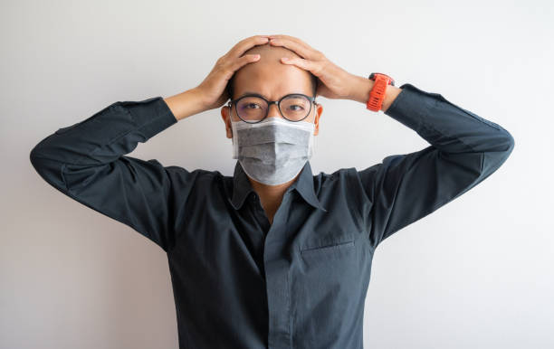 asiatische geschäftsmann mit kopfschmerzen und gestresst, während das tragen chirurgische maske den ganzen tag in covid-19 pandemie. - skinhead stock-fotos und bilder