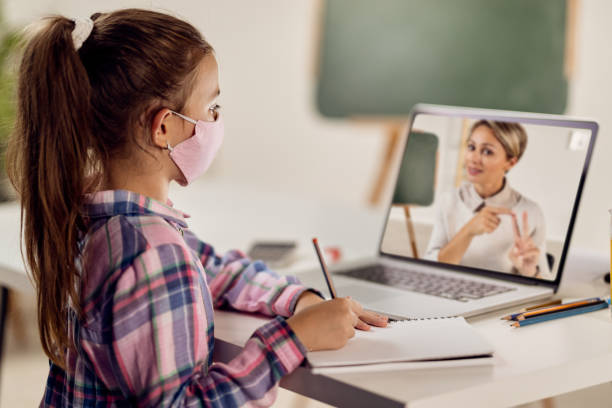 Schoolgirl writing while having online class with her teacher over laptop due to virus pandemic. Little girl wit face mask using laptop and writing in notebook while following online class during COVID-19 lockdown. lifestyles teaching little girls child stock pictures, royalty-free photos & images