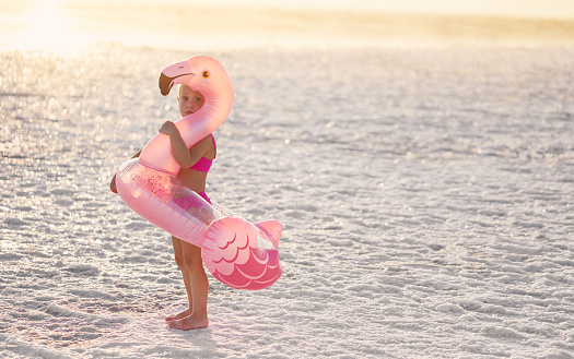 The end of the summer. Cute sad  child girl in a pink swimsuit holds an inflatable pink flamingo on the tropical beach. Place for text\