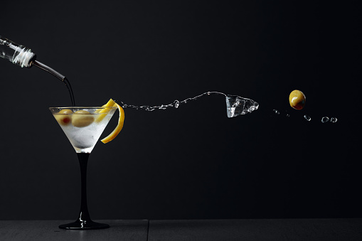 Martini with green olives, ice and lemon peel. Cocktail with splashes on a black background. Copy space.