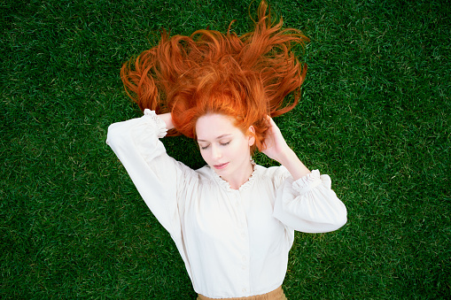Redhead woman is resting on bright green lawn, her eyes closed and dreaming. Model with long thick hair enjoys summer