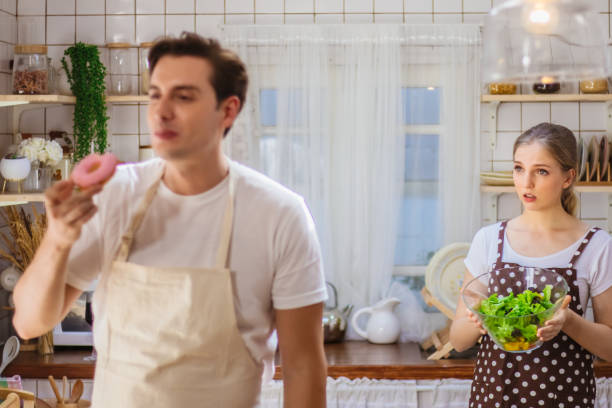 beau couple caucasien ou amant dans la cuisine ensemble. l’homme heureux mangent la malbouffe malsaine comme donut tandis que la salade de cuisson de femme pour le repas. petite amie malheureuse bouleversée ou déçue sur de mauvaises habitudes de petit - overweight women salad frustration photos et images de collection