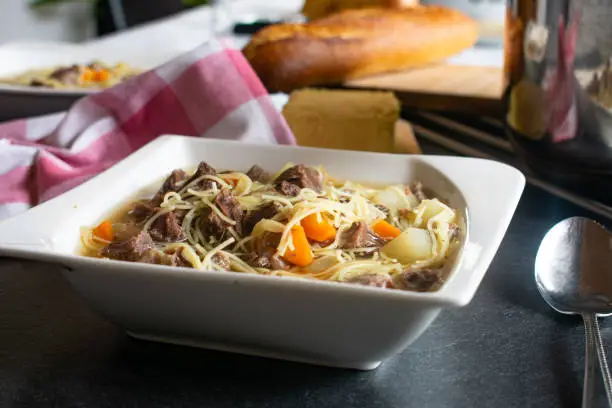 fresh and homemade cooked noodle soup with tender beef meat on a white plate served with bread and butter on a table - ready to eat food