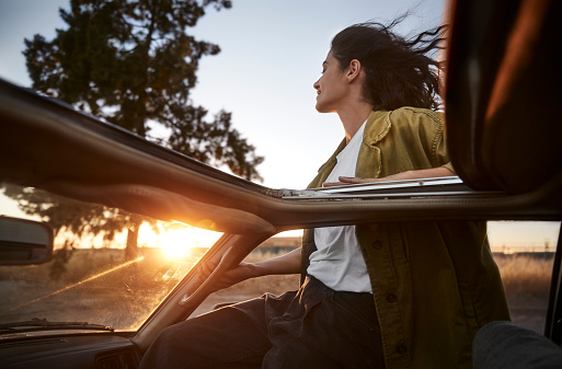 Happy young latina hipster woman leaning out of car window sitting on car door at sunset feeling wind in her face