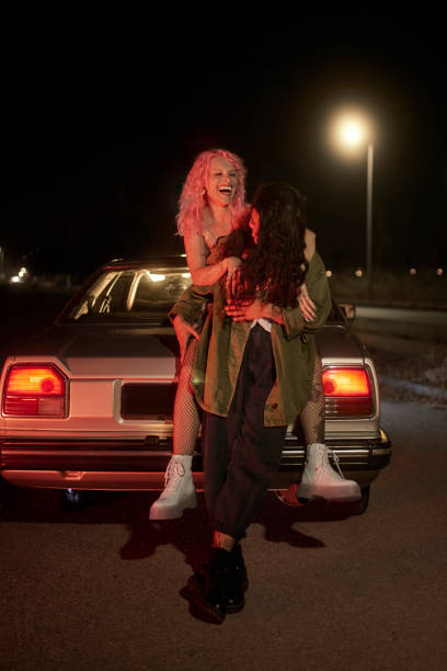 Two girl friends leaning on car having fun at night Two hipster alternative girl friends leaning on car having fun at night alternative lifestyle photos stock pictures, royalty-free photos & images