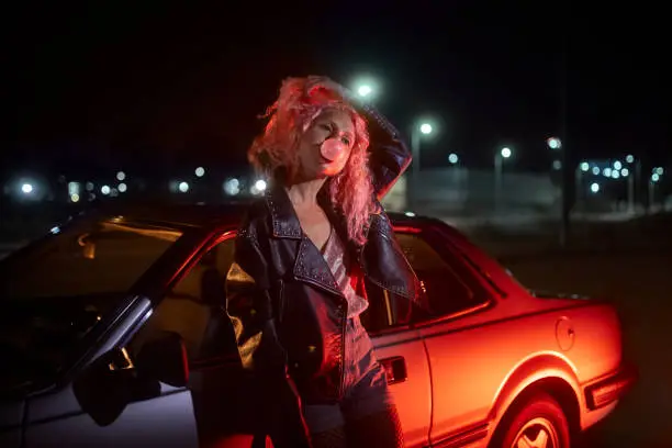 Alternative young hipster girl leaning on car with bubble gum at night