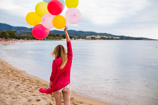 Young casually dressed woman carrying colorful balloons walking at the beach on a sunny summer day in Greece