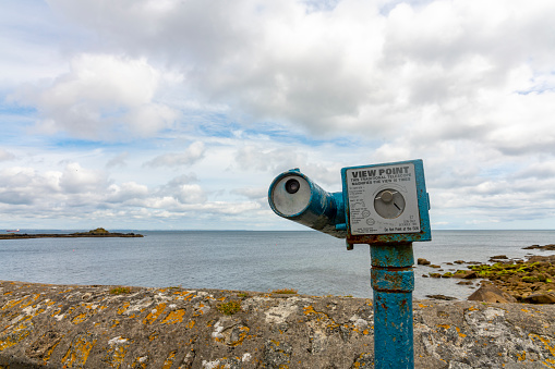 A pair of shiny gold metal or brass binoculars fixed to a tripod and pointed to observe the view of the pacific ocean and blue sky on a sunny day. The binoculars point in the direction of the sea ocean waterfront across the view of blurred grass and a hedge in a garden backyard.