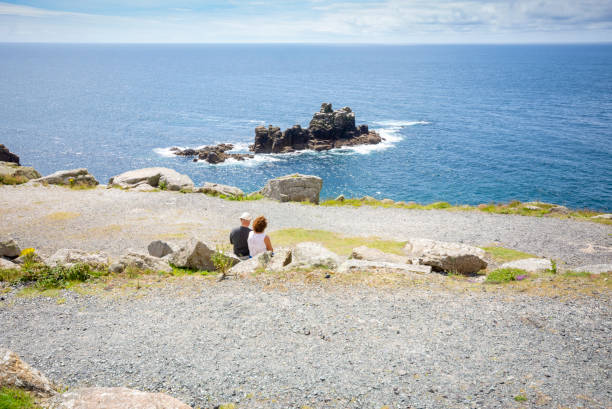 View from the Land's end peninsular, Cornwall, England, UK, the most westerly point on the English mainland. This shows the rocks off the coast with a senior couple sat looking at the view.