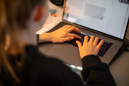 An over the shoulder view of a teenage girl working on a laptop in a home office.