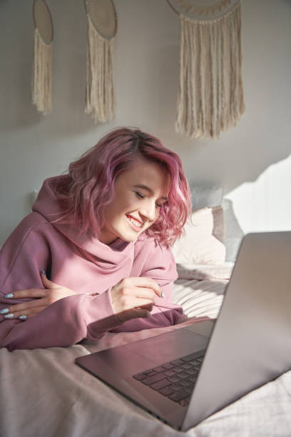 счастливый хипстер gen z подросток девушка с розовыми волосами с помощью ноутбука в постели. вертикальной - gen z стоковые фото и изображения