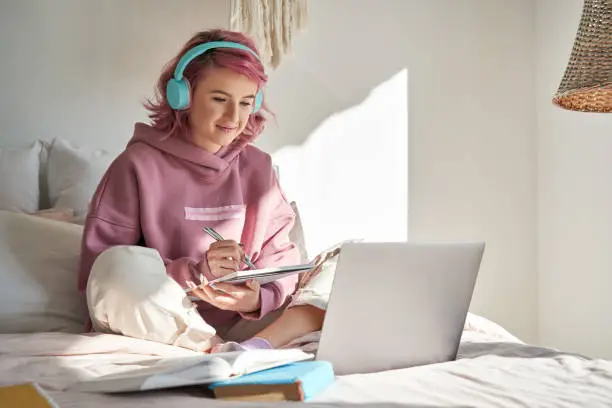 Photo of Hipster teen girl student with pink hair watch online webinar learning in bed.