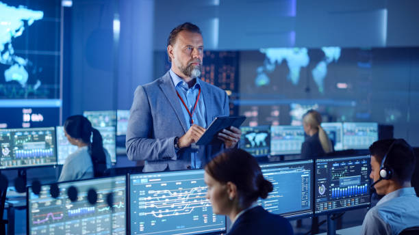 istock Male Project Leader is Standing with Tablet Computer and Making Mental Calculations. Data Science Engineers Work Around Him. Telecommunications Control Monitoring Room with a Global Map on Big Screen. 1266953176