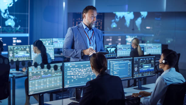 istock Male Project Leader with Tablet Computer Talks to Young Data Science Engineers and Checks Up on Their Work Progress. Telecommunications Control Monitoring Room with a Global Map on Big Screen. 1266953184