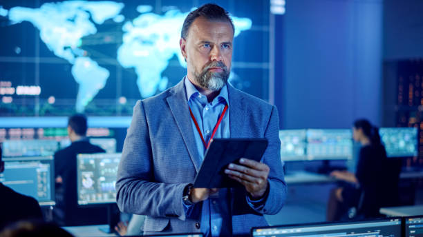 istock Male Project Leader is Standing with Tablet Computer and Making Mental Calculations. Data Science Engineers Work Around Him. Telecommunications Control Monitoring Room with a Global Map on Big Screen. 1266953209