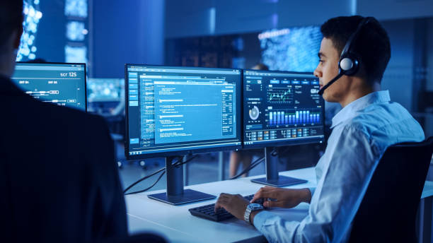 istock Confident Male Data Scientist Works on Personal Computer Wearing a Headset in Big Infrastructure Control and Monitoring Room. Young Engineer in a Call Center Office Room with Colleagues. 1266953216