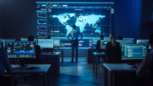 istock Project manager Stands Next to Big Screen with Neural Network. Team of Professional Computer Data Science Engineers Work on Desktops in a Dark System Control and Monitoring Telecommunications Office. 1266953242